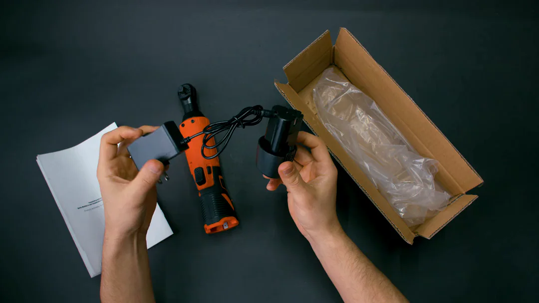 Top-down: hands hold the wall charger and the 12V battery with its charging cable plugged in, beside the SuperRatchet, the instruction sheet and the open carton